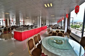 A spacious dining area with round tables and chairs, a central buffet, and large windows offering a view.