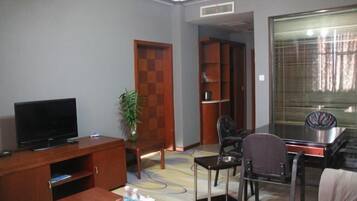 A compact living room with a flat-screen TV, a wooden cabinet, a dining table with chairs, a potted plant, and a large window with blinds.