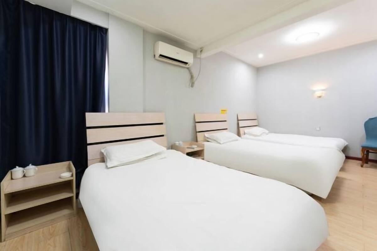 A hotel room with two single beds, a small bedside table with a lamp, a chair, and an air conditioning unit on the wall.