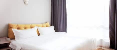 A neatly made bed with white bedding and a mustard-colored headboard, a bedside table, and a window with sheer curtains.