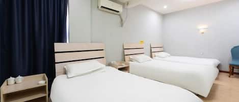 A hotel room with two single beds, a small bedside table with a lamp, a wall-mounted air conditioner, and a window with dark curtains.