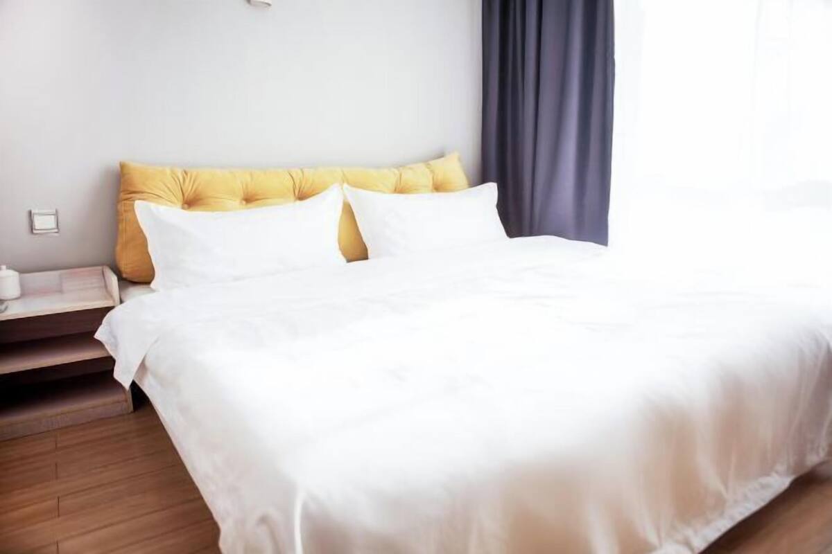 A neatly made bed with white bedding and two yellow pillows, a bedside table with a white lamp, and a dark curtain to the right.