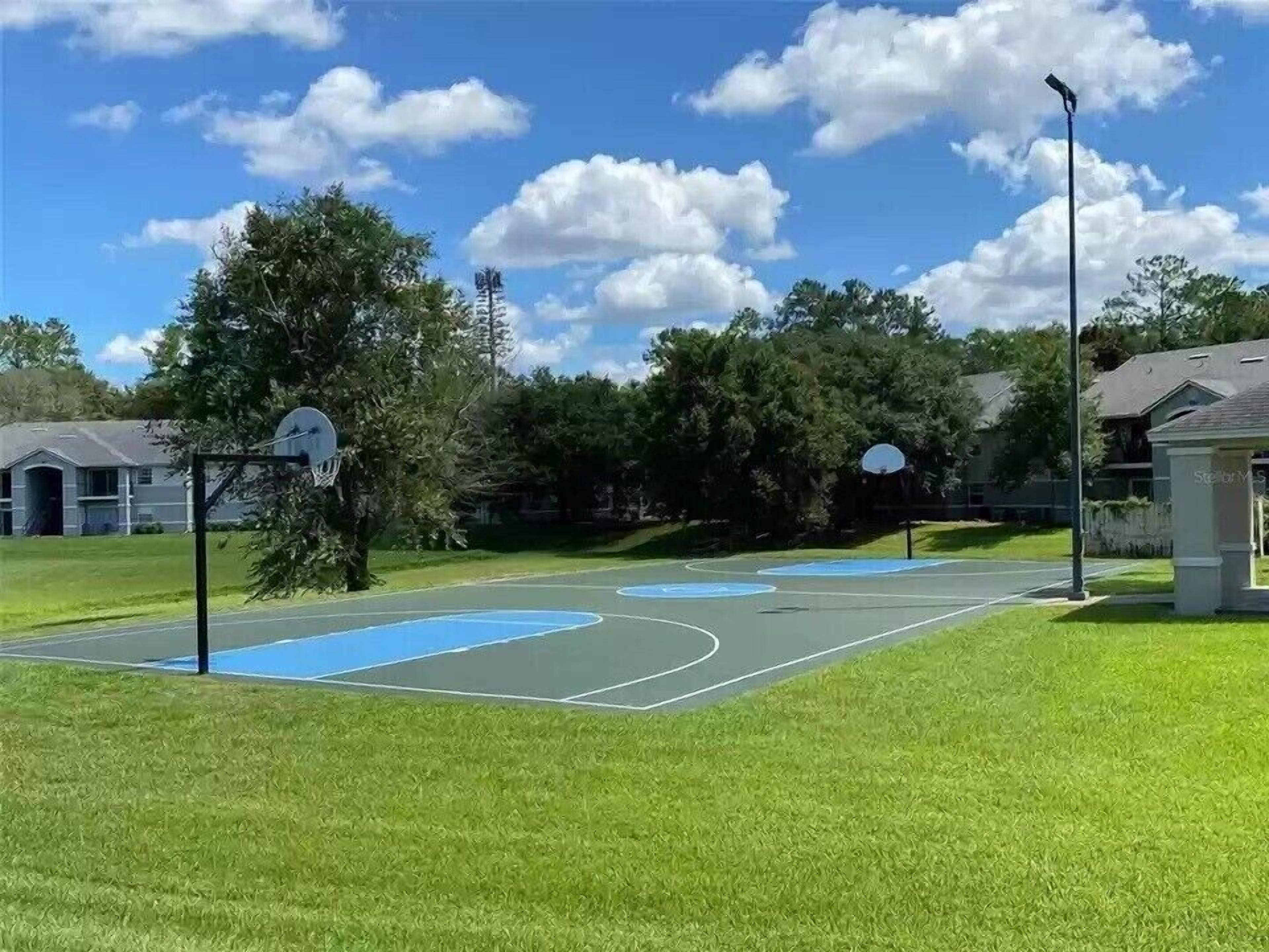An outdoor basketball court with two hoops, surrounded by trees and a clear sky.