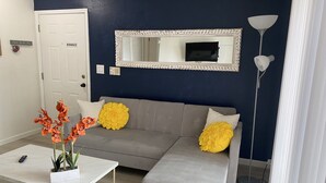 A living room with a grey sofa, a white coffee table, and a potted plant with orange flowers. - Botanical Retreat: King Comfort & Poolside Peace (Gainesville)