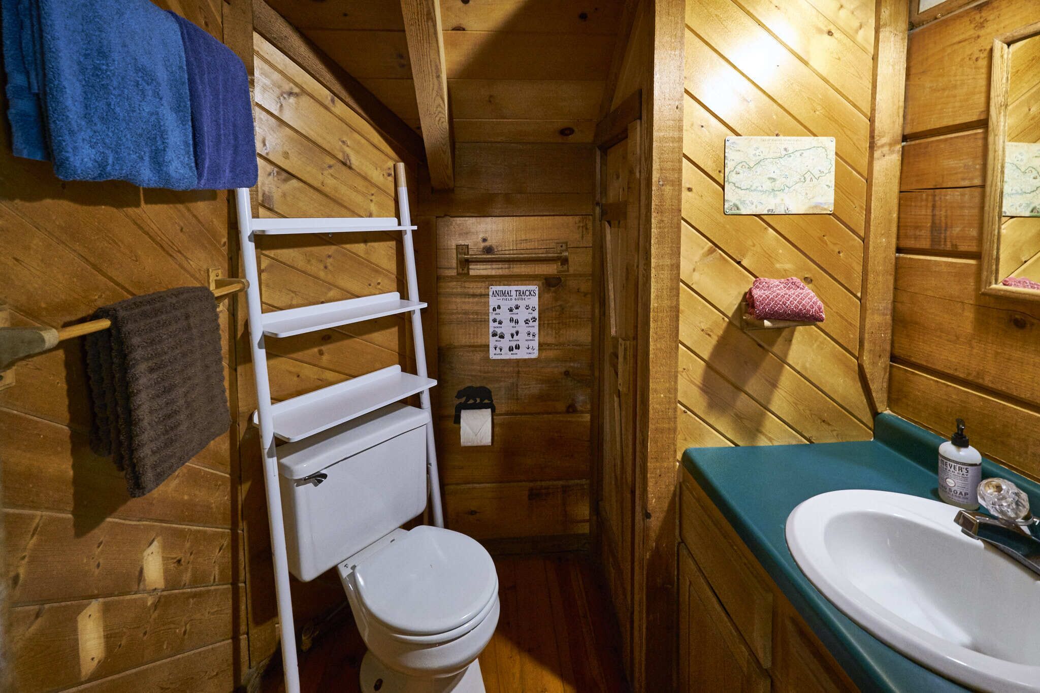 A small, wooden cabin bathroom with a white toilet, a shelf with towels, a sink with a mirror, and a sign indicating animal tracks.