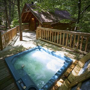 Un jacuzzi en una terraza de madera rodeado de árboles, con una cabaña de madera al fondo.