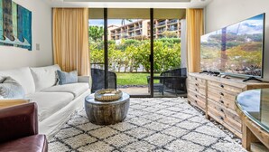 Ein modernes Wohnzimmer mit Sofa, Couchtisch, Fernseher und Blick auf ein Gebäude sowie Grünflächen durch große Fenster.