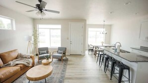 A spacious living room with a brown leather sofa, a round wooden coffee table, and a grey armchair. A ceiling fan is visible, and there is a large window providing natural light. - The Sandbar #41 - Lake, Pool and Hot Tub Access! (Garden City)