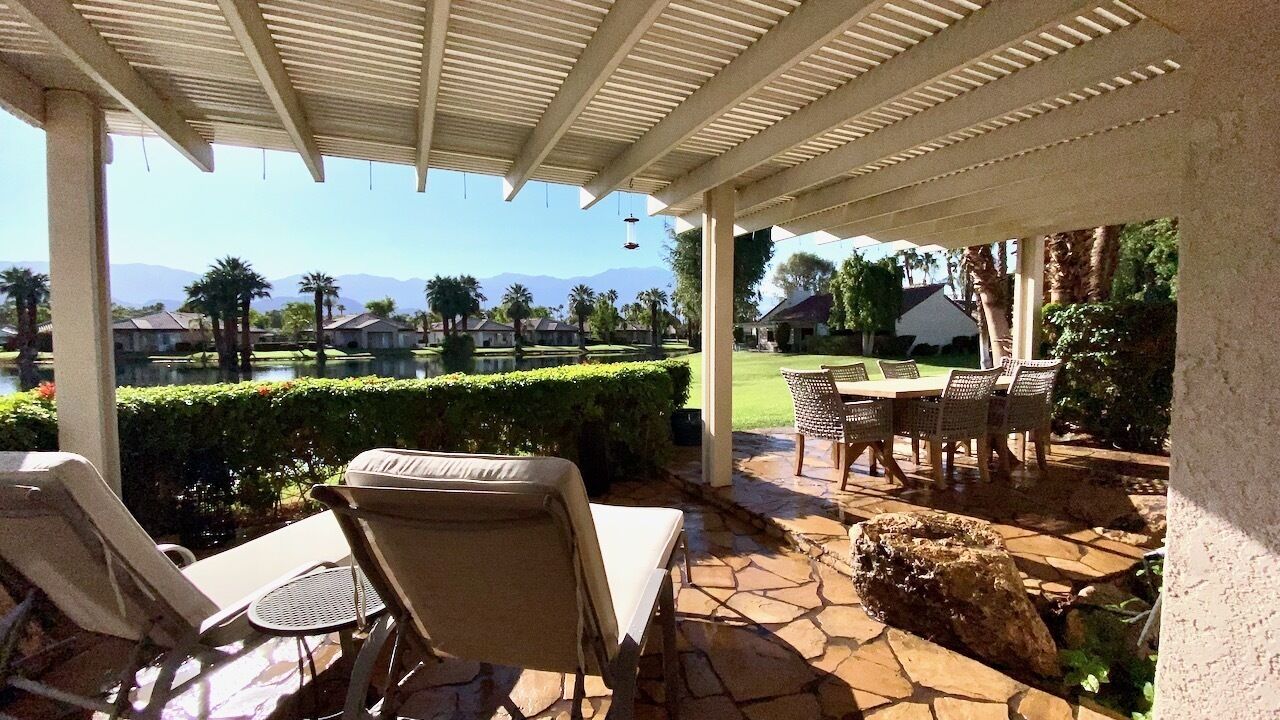 Un patio techado con mobiliario exterior, piso de piedra y vistas a un jardín ajardinado y casas a lo lejos.