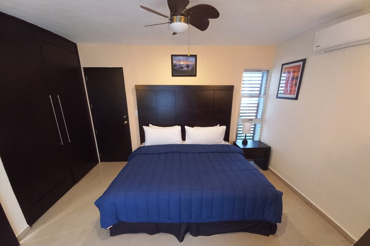 A bedroom with a large bed, a ceiling fan, a dark headboard, a nightstand with a lamp, and a window with blinds.