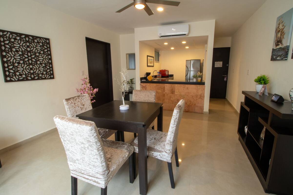 A dining area with a table and chairs, a kitchen in the background, and a ceiling fan.