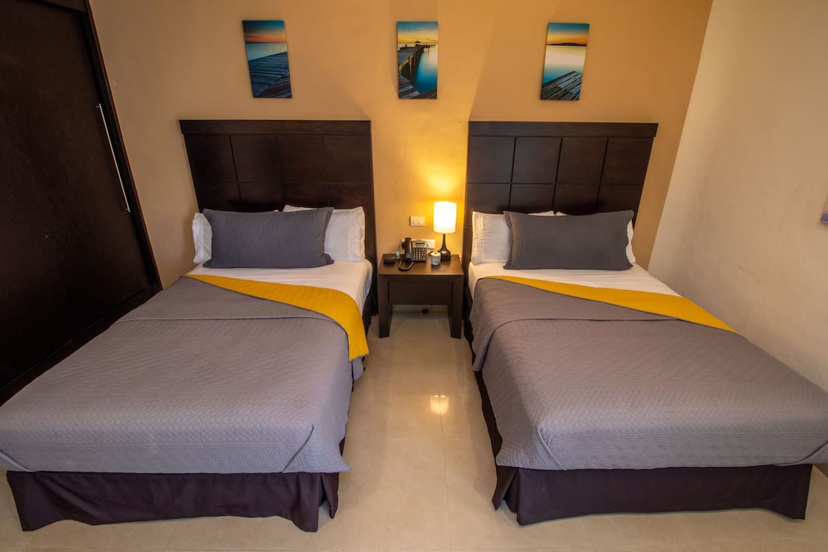 A hotel room with two beds, each with a headboard, a bedside table with a lamp, and artwork on the wall.