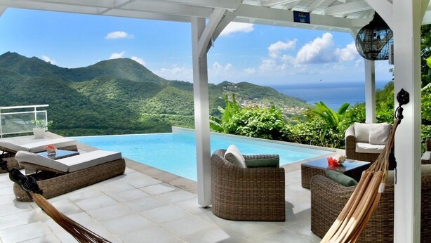 A covered outdoor area with a pool, wicker furniture, and a view of mountains and the sea.