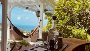 A wooden table with a bottle of liquor, a glass, and a knife holder. A hammock is visible in the background, and a view of the ocean is seen through a white structure.