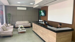 A modern reception area with a wooden reception desk, a flat-screen TV mounted on the wall, a sofa, a coffee table with flowers, and a potted plant. - HOTEL SEVEN (Goiânia)