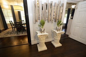 Un pasillo con piso de madera oscura, un comedor con mesa y sillas, y una pared con tablones de madera verticales y dos pedestales blancos con plantas.