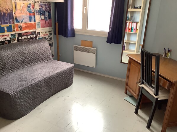 Une pièce avec un canapé gris, un bureau en bois avec une chaise, un radiateur et une fenêtre avec des rideaux.