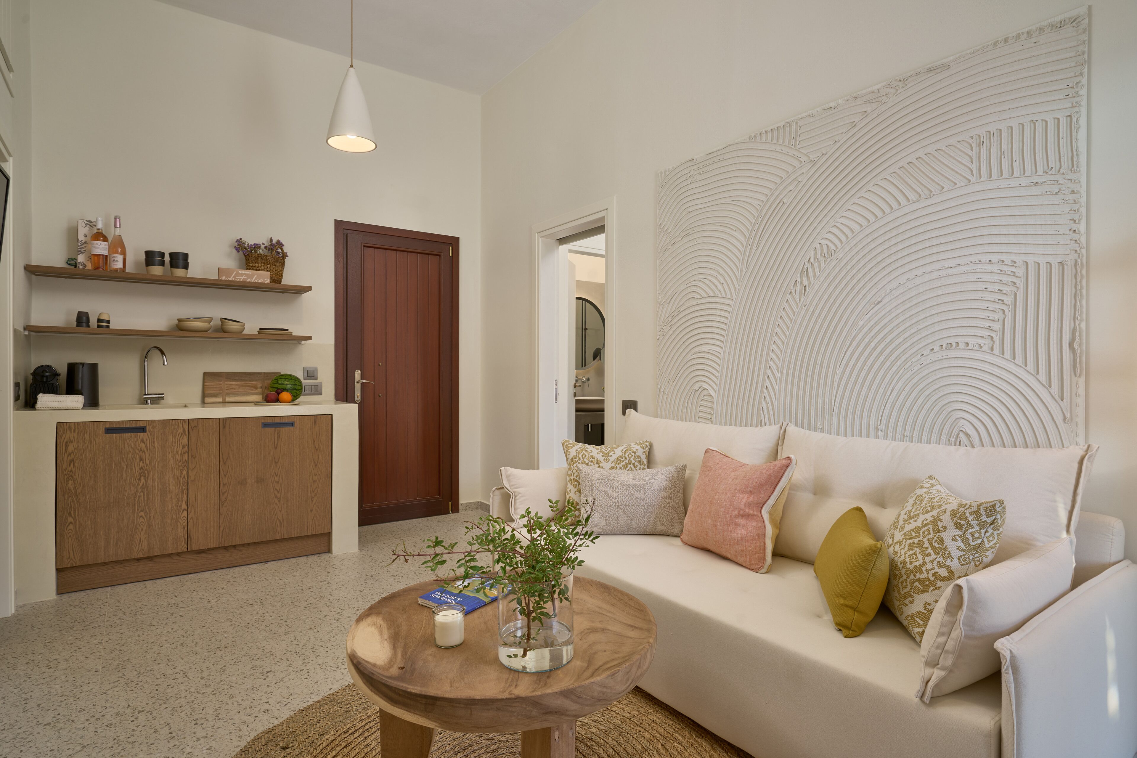 A modern living room with a wooden coffee table, a white sofa with patterned cushions, and a kitchen area with wooden cabinets and shelves.