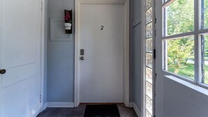 A hallway with a white door marked with the number 2, a small potted plant on the wall, and a window with a view of greenery. - Eclectic Escape #2 - Team Joseph Ellen (Jacksonville)