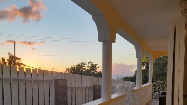 A covered outdoor seating area with a white railing, a green cushioned bench, and a view of a wooden fence and trees under a sunset sky. - La Kayé Celesté your home away from home!! (Castries)