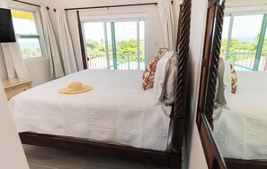 A bedroom with a large bed, a view of greenery through the window, and a wooden headboard.