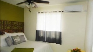 A bedroom with a bed, a ceiling fan, an air conditioning unit, a window with curtains, and a bedside table with a flower arrangement. - La Kayé Celesté your home away from home!! (Castries)