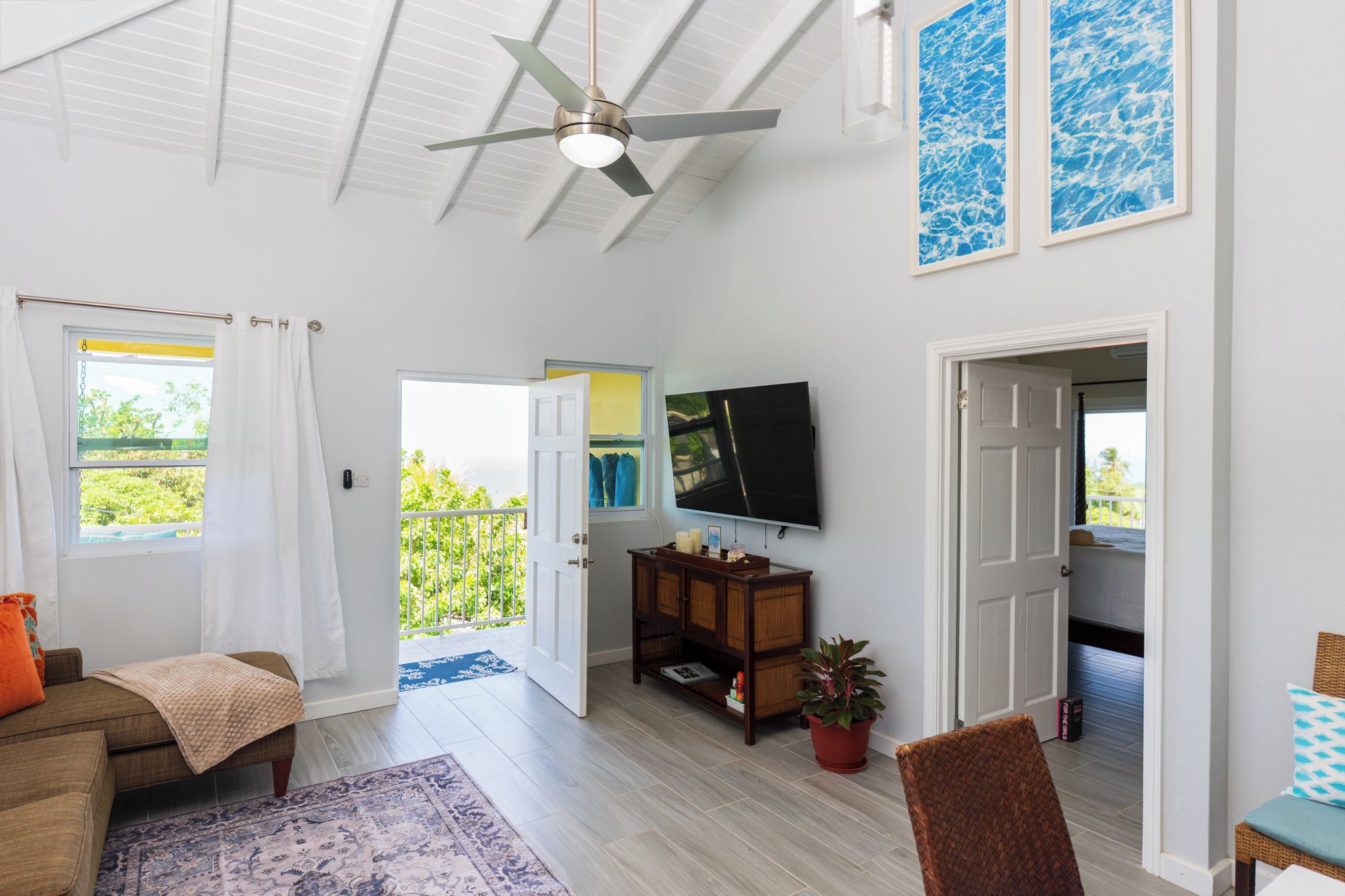A spacious living room with a ceiling fan, a flat-screen TV mounted on the wall, and a large window offering a view of the outdoors.