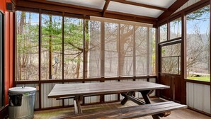Un patio cubierto con una mesa y bancos de madera, ventanas amplias con vistas a árboles, y un bote de basura metálico.