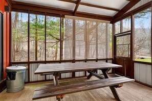 Une terrasse couverte avec une table et des bancs en bois, de grandes fenêtres donnant sur les arbres et une poubelle en métal.