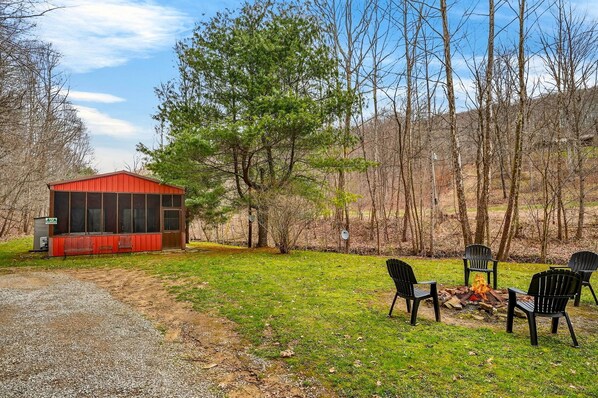 A red cabin with a screened porch, surrounded by a grassy area with a fire pit and chairs.
