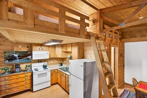 A wooden cabin interior with a kitchen, wooden beams, a stove, a refrigerator, and a television. - Lazy Lane Cabins - Creekside (Laurelville)