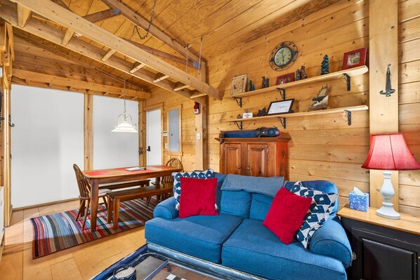 A cozy cabin interior with a wooden ceiling, a dining area, a blue sofa with red cushions, and a mounted clock.