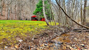A forested area with a small red building, a stream, and a grassy field. - Lazy Lane Cabins - Creekside (Laurelville)