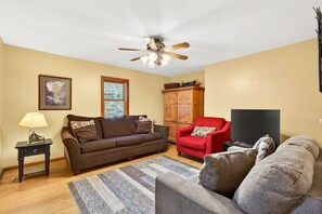 Un salon avec un canapé marron, un fauteuil rouge, un meuble en bois, une télévision et un ventilateur de plafond.