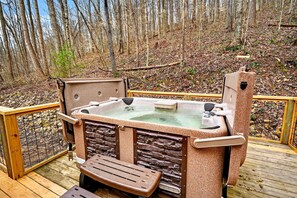 Un bain à remous sur une terrasse en bois agrémentée d’éléments en pierre, entouré d’une balustrade en bois et d’une zone boisée.