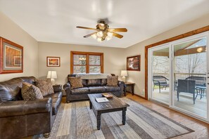 A living room with a leather sofa, a coffee table, and a ceiling fan. There are framed pictures on the walls and a large window with a view.