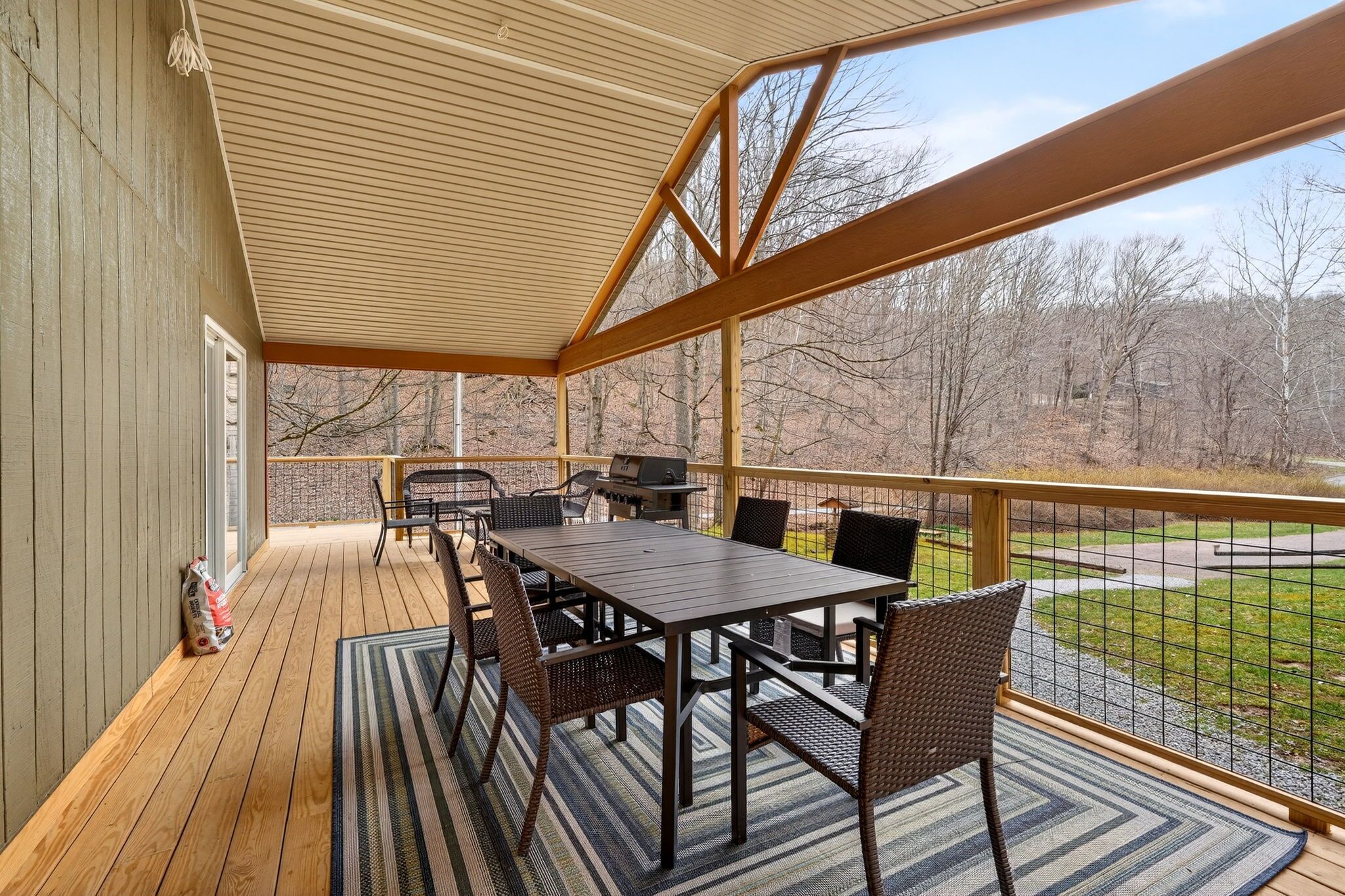 A covered outdoor deck with a table and chairs, a grill, and a view of a wooded area.