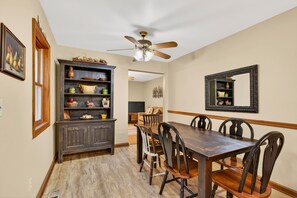 A dining room with a wooden table, chairs, a ceiling fan, a dark wooden cabinet, and a mirror on the wall.