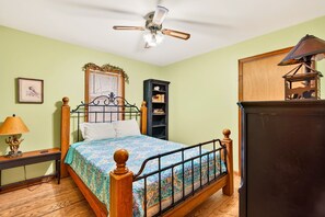 Een slaapkamer met een bed, plafondventilator, nachtkastje, lamp, een boekenplank en een spiegel.