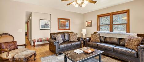 A living room with a brown leather sofa set, a wooden coffee table, a ceiling fan, and a large window with a view.