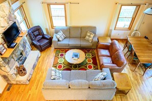A living room with a stone fireplace, a grey sofa set, a brown leather armchair, a wooden dining table, and a colorful rug.