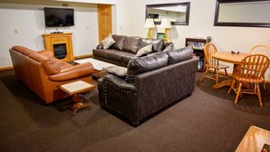 Ein Wohnzimmer mit braunem Ledersofa, einem dunkel gemusterten Sofa, einem Holztisch, einem Esstisch mit Holzstühlen, einem an der Wand befestigten Fernseher und einem Kamin.