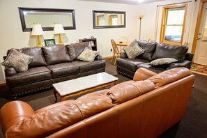 A living room with two brown leather sofas, a coffee table, and a bookshelf. - Lazy Lane Cabins - Tree-Top Chalet (Logan)