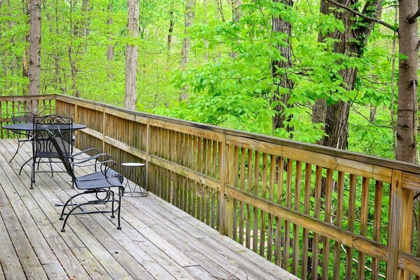 A wooden deck with metal chairs and tables, surrounded by a wooden railing, overlooking a forest.