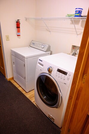 A laundry room with two washing machines, a fire extinguisher, and a shelf with cleaning supplies.