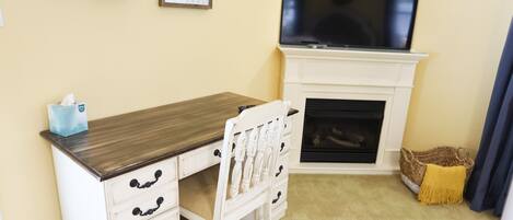 A room with a wooden desk, white drawers, a television above a fireplace, and a basket by the window.