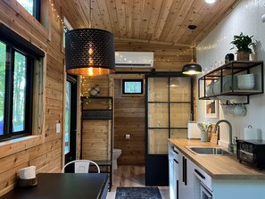 A compact kitchen with wooden walls, a black hanging light fixture, a window, and a sink. - The Dogwood Tiny House (Logan)