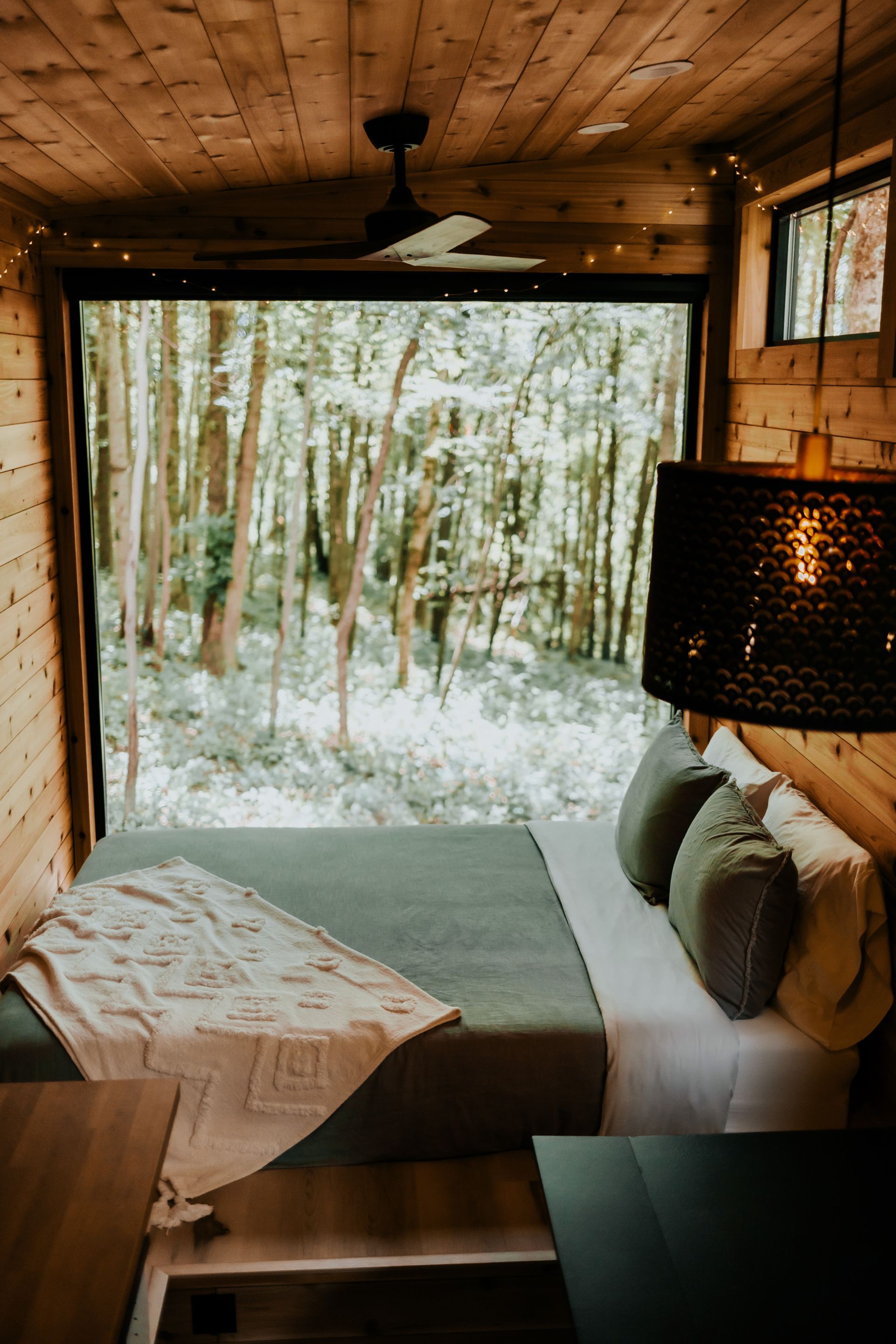 A cozy cabin interior with a bed, pillows, a table, and a view of a forest through a large window.