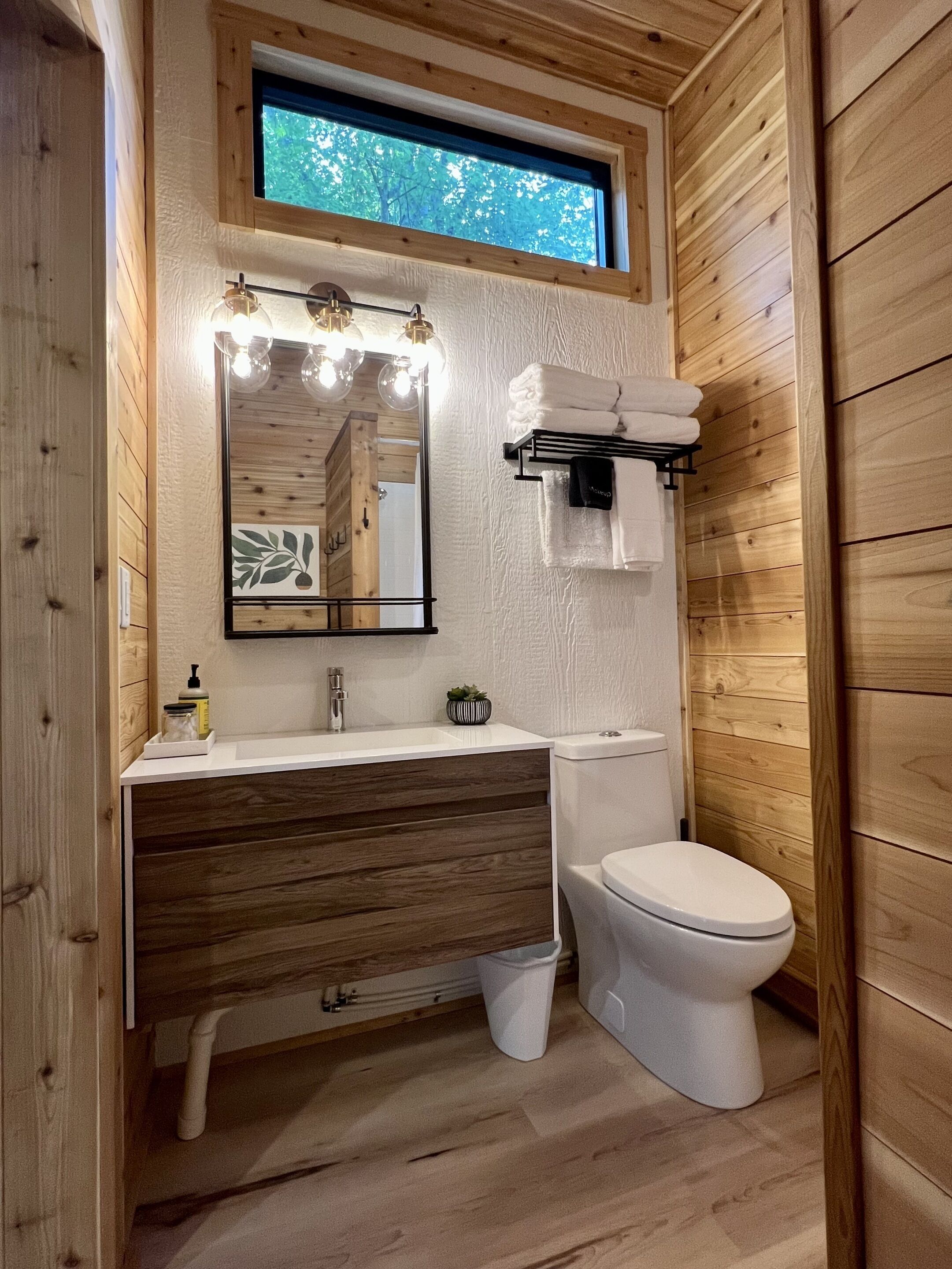 A compact bathroom with a wooden interior, a white sink, a mirror with lights, a toilet, and a window above the sink.