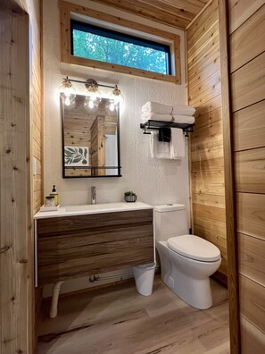 A compact bathroom with a wooden interior, a white sink, a mirror with lights, a toilet, and a window above the sink. - The Dogwood Tiny House (Logan)